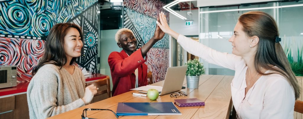Group of positive young multiethnic female colleagues smiling and giving high five while sitting at wooden table with documents and laptop in creative workspace